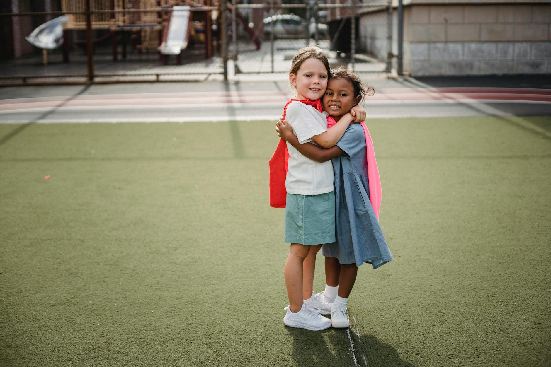 Two young girls wearing capes joyfully embrace on a playground, symbolizing friendship and fun.